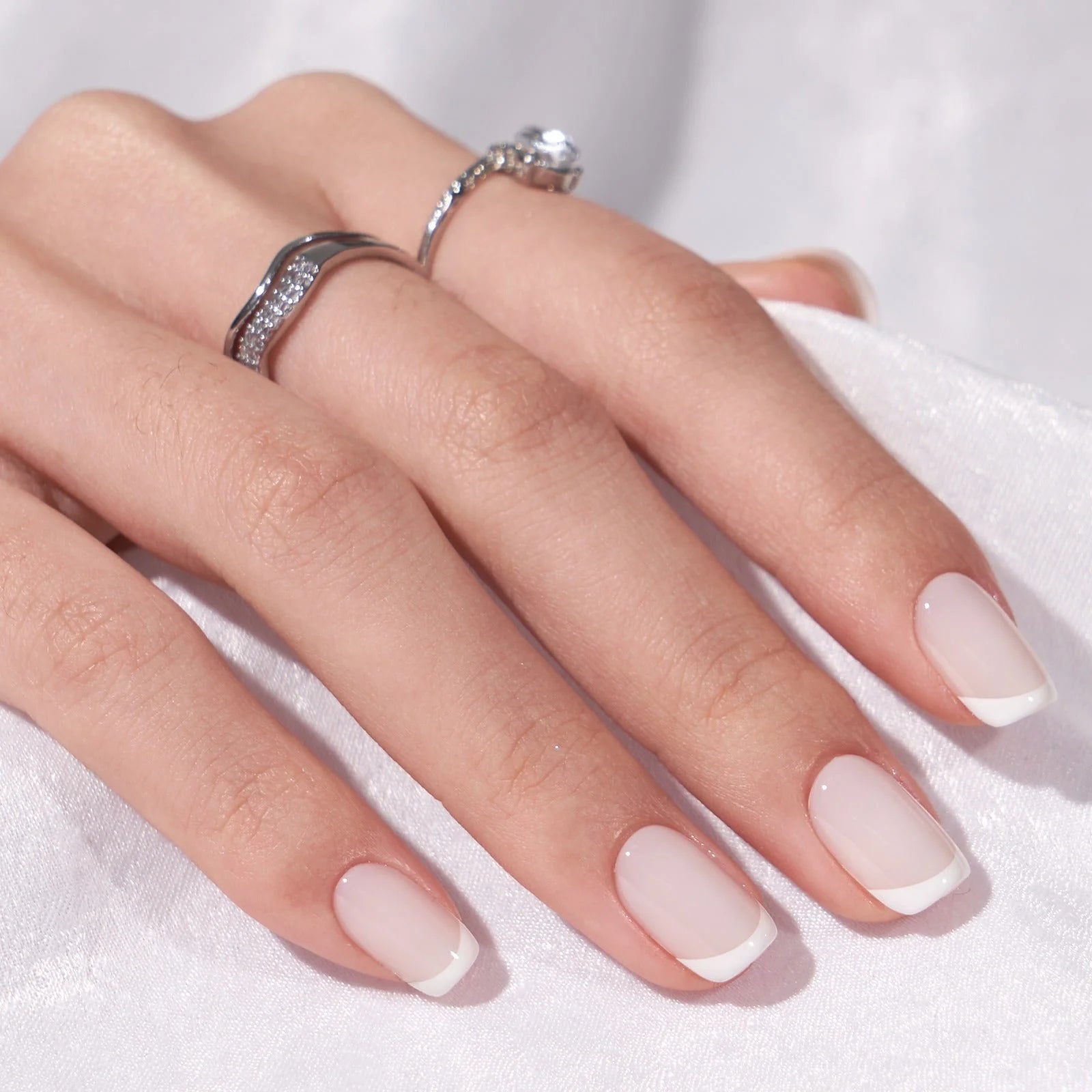 Frosted French gelénaglar närbild – square fransk manikyr med silverringar mot vitt tyg