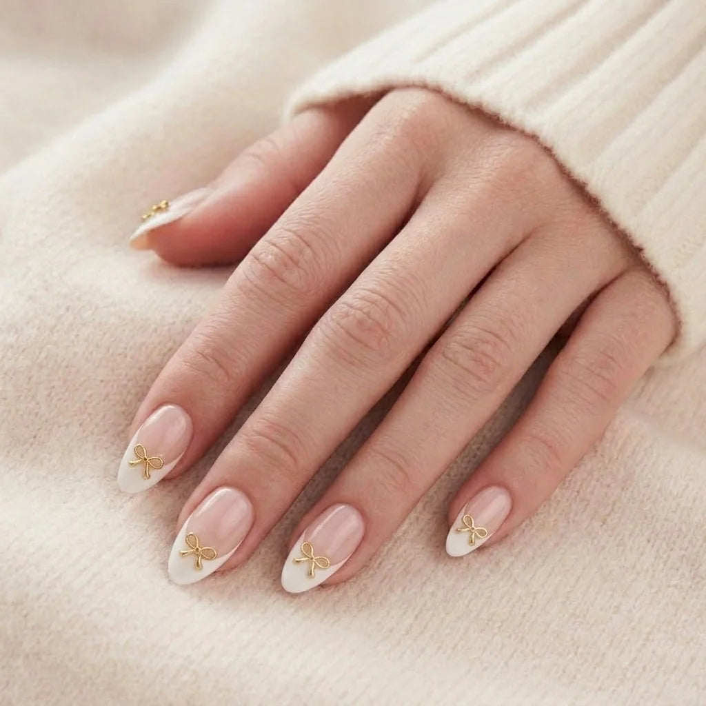Blushing Bow naglar på hand – mandelformade lösnaglar med fransk vit tipp och 3D guldrosett mot beige stickad tröja