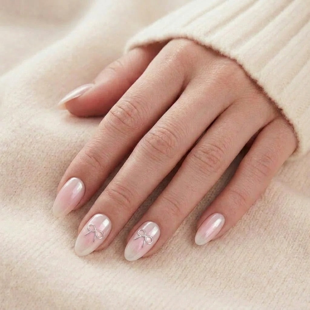 Aura Amoré faux ongles en chrome nacré avec ombré rose blush et nœud argenté sur faux ongles amande, devant un pull tricoté beige