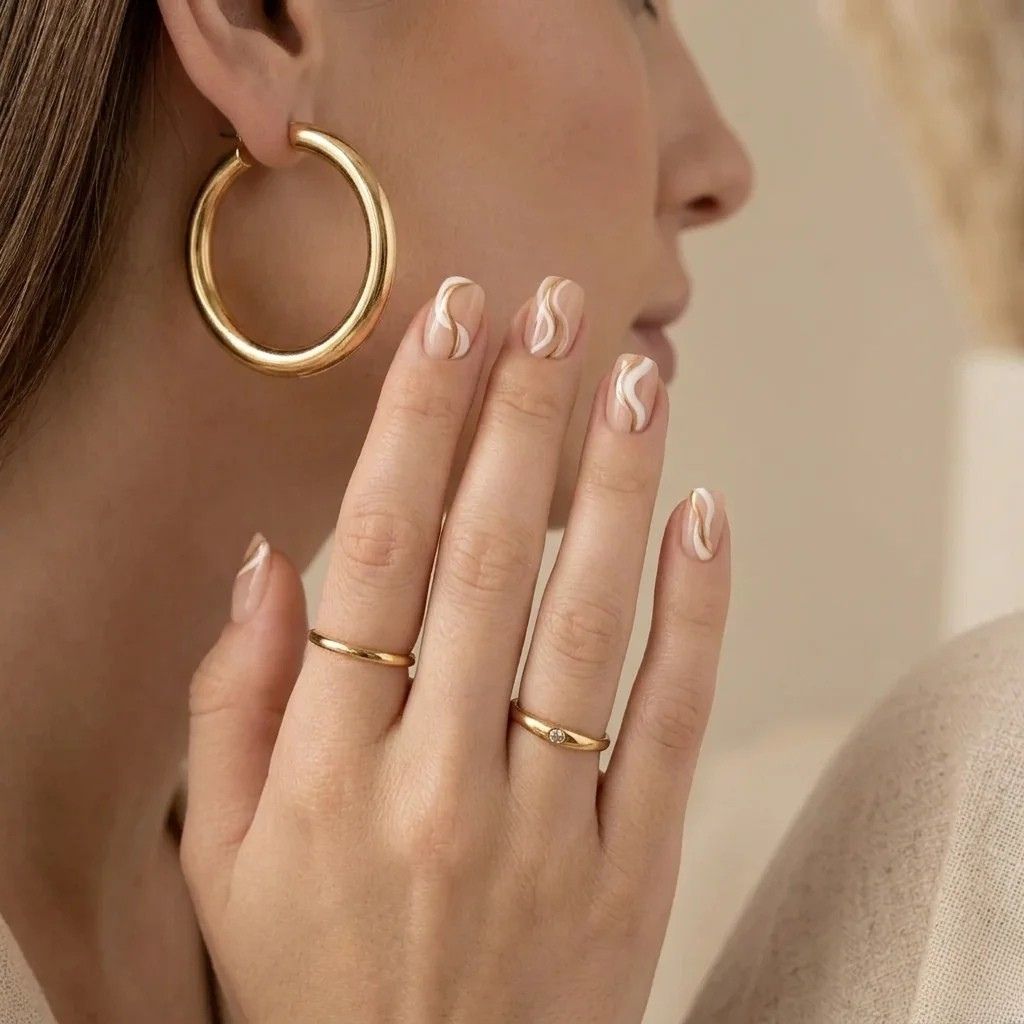 Faux ongles courts et arrondis nude à lignes ondulées blanches et beiges. Main aux bagues en or.